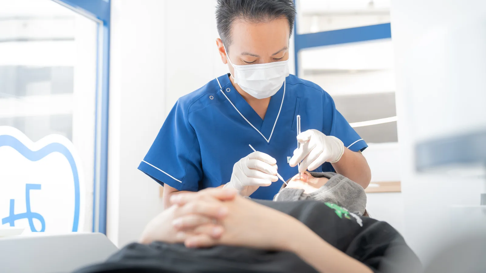 青いスクラブとマスクを着けた歯科医師が患者の口腔内を器具で診察している治療風景