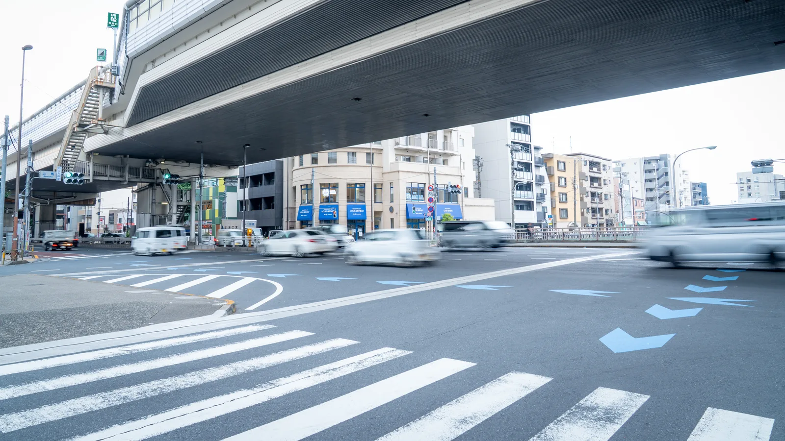 高架道路の下を車が行き交う交差点で、横断歩道と青い矢印標示が見える街並みの様子